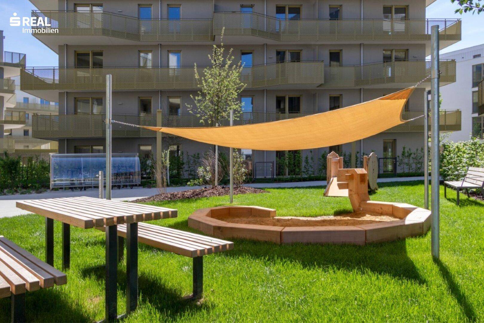 Kinderspielplatz mit Sandkasten, Picknicktisch und Sonnensegel in einer grünen Umgebung.