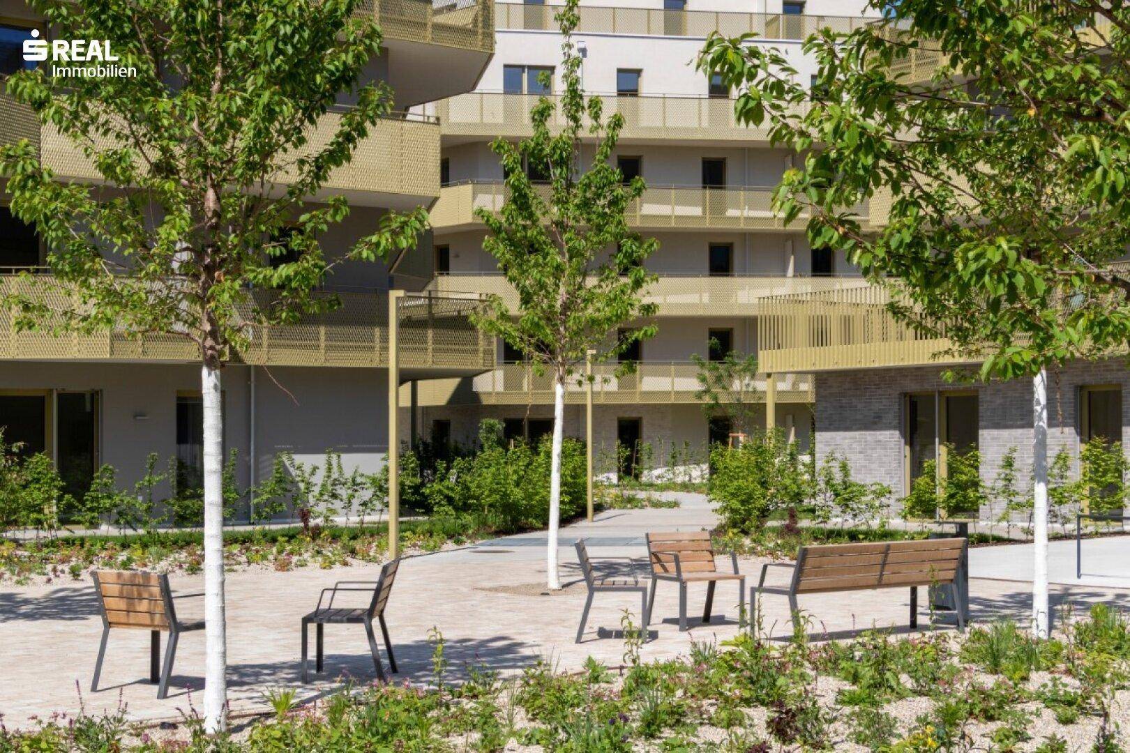 Gemeinschaftlicher Gartenbereich mit Sitzgelegenheiten und jungen Bäumen zwischen den modernen Wohngebäuden.