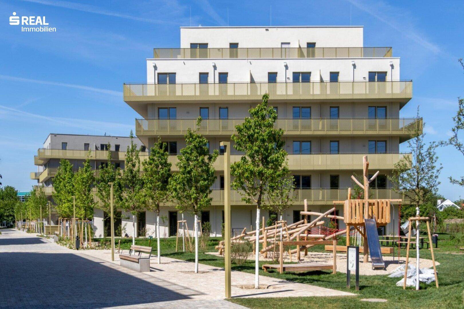Moderner Kinderspielplatz mit Holzgeräten und Grünflächen vor einer Wohnanlage.