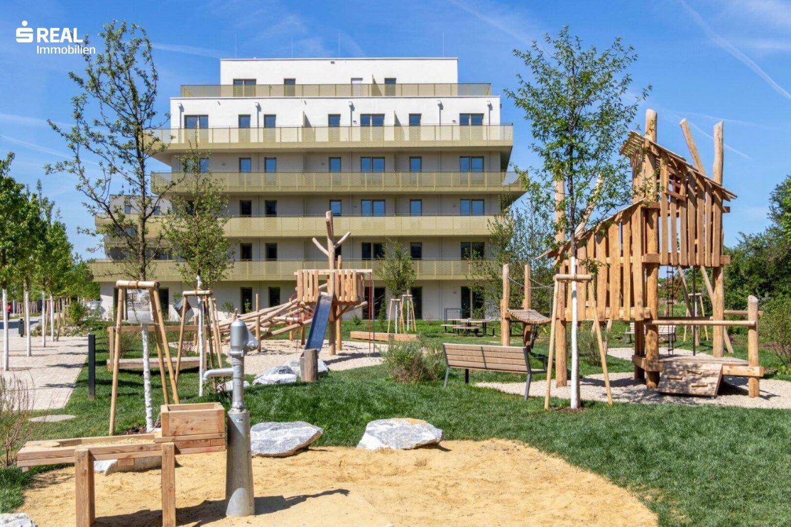 Neubauwohnanlage mit großem Kinderspielplatz und modernen Gebäuden unter strahlend blauem Himmel.