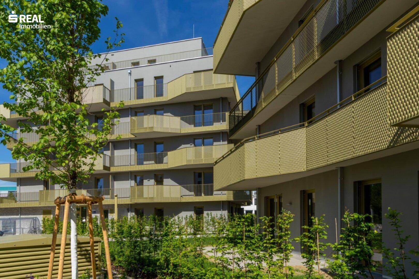 Seitenansicht eines neuen Wohngebäudes mit mehreren Balkonen und jungen Bäumen.