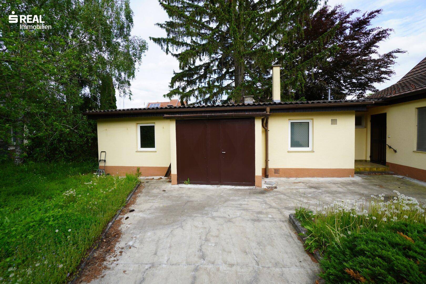 Ansicht der Garage mit braunem Tor und angrenzendem Gebäude, umgeben von Grün.