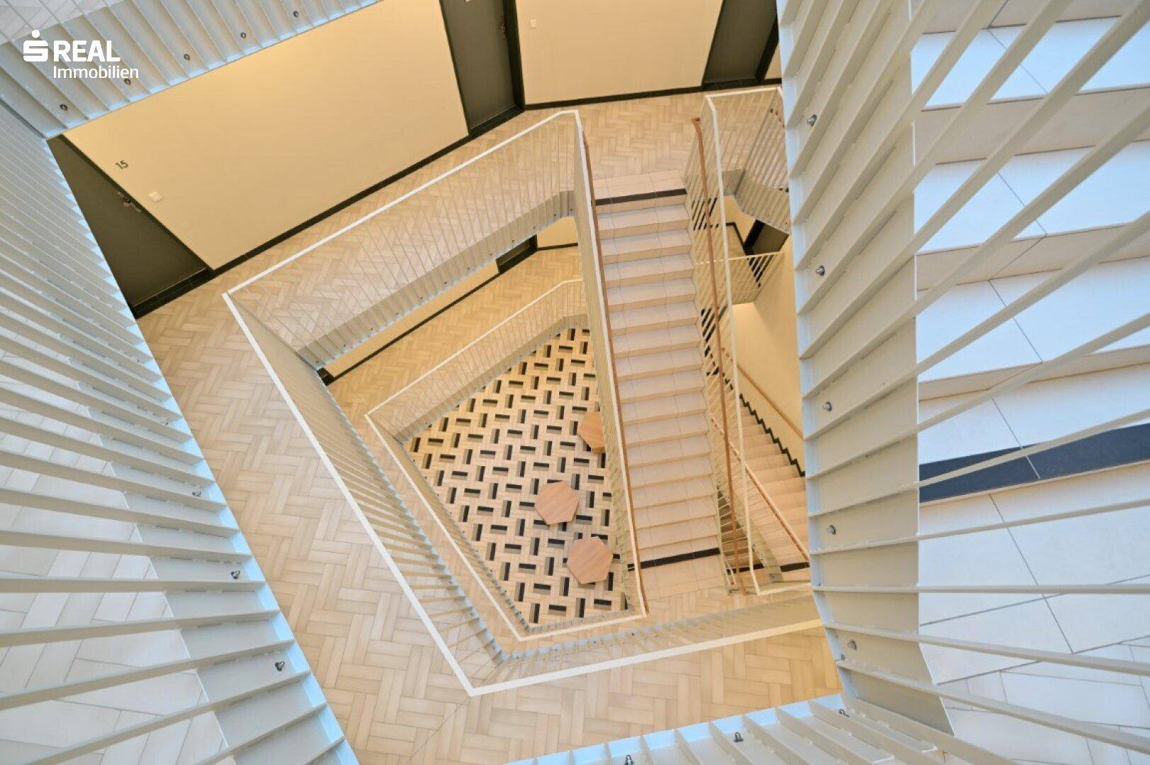 Blick von oben in ein modernes Treppenhaus mit geometrischem Bodenmuster und mehreren Etagen.