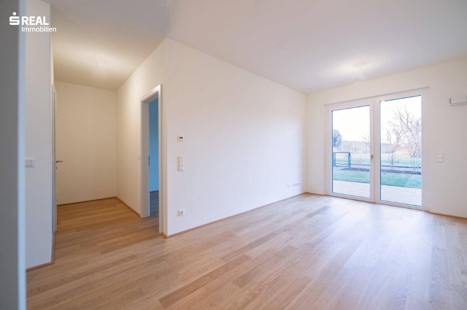 Heller, leerer Raum mit Holzboden und großer Glastür, die zu einem Gartenbereich führt.