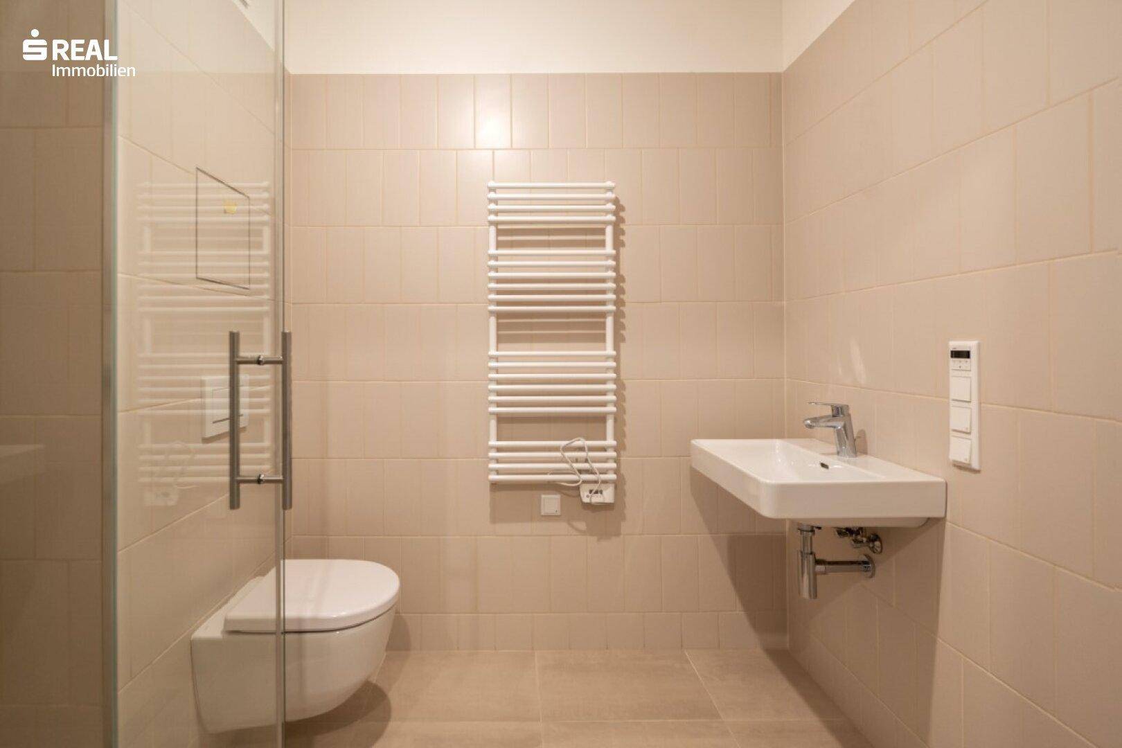Modernes Badezimmer mit beigefarbenen Fliesen, wandhängendem WC, Einzelwaschbecken und Handtuchheizkörper.