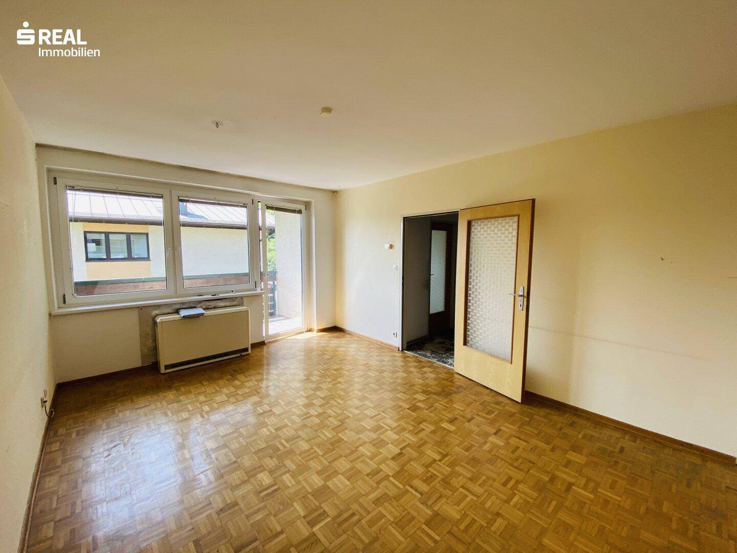 Geräumiges Wohnzimmer mit Parkettboden, großen Fenstern und Zugang zum Balkon.