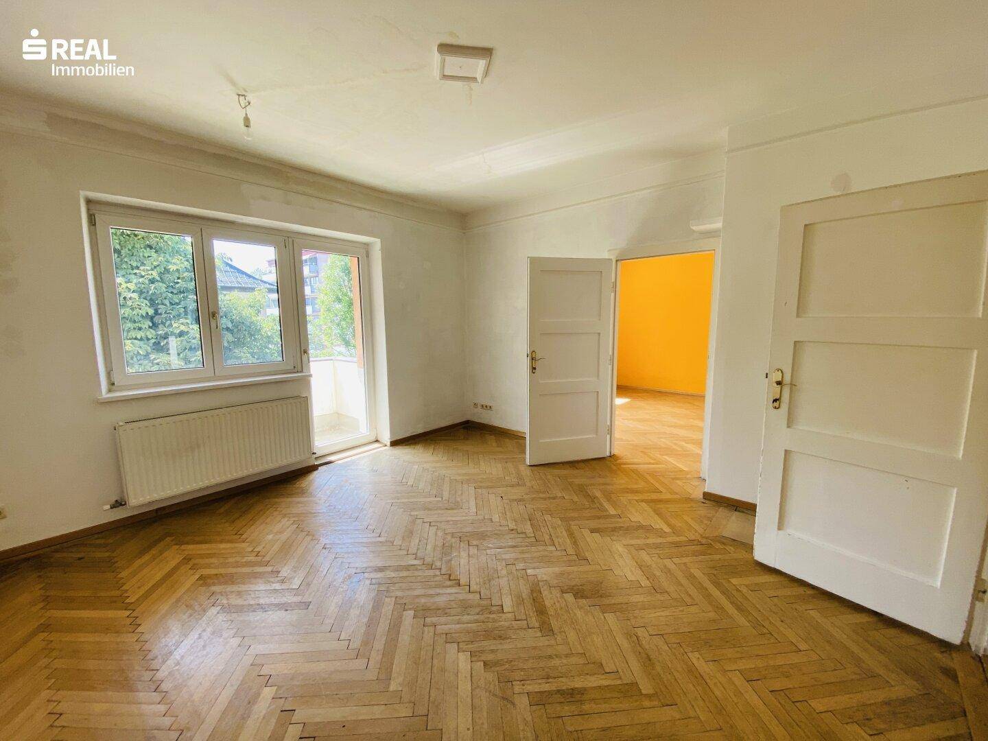Helles Wohnzimmer mit Fischgrätparkettboden, Zugang zum Balkon und Blick ins Grüne.