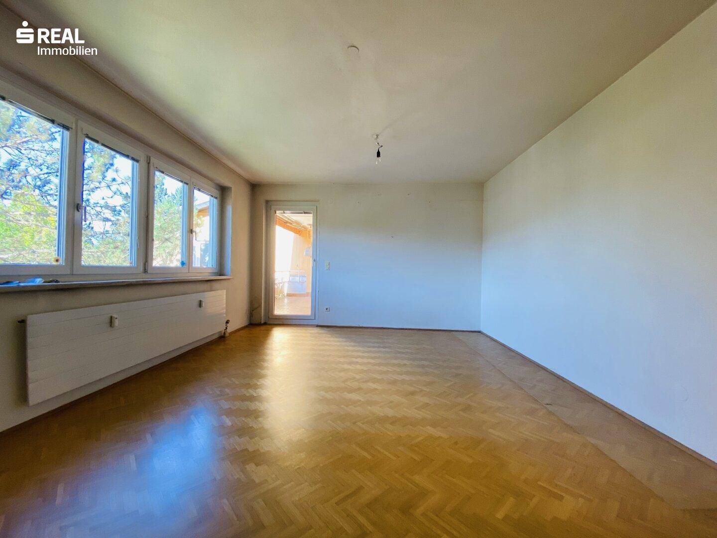 Helles, leeres Wohnzimmer mit Fischgrätparkettboden und Zugang zu einem Balkon.