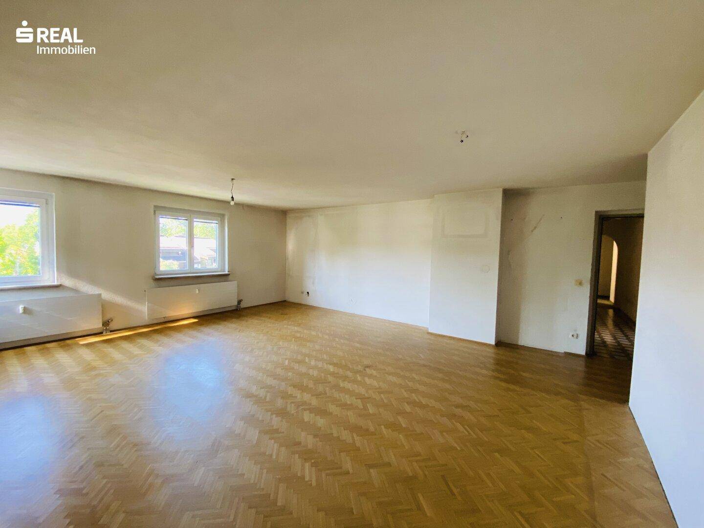 Geräumiges, leeres Wohnzimmer mit Parkettboden und mehreren Fenstern für viel Tageslicht.