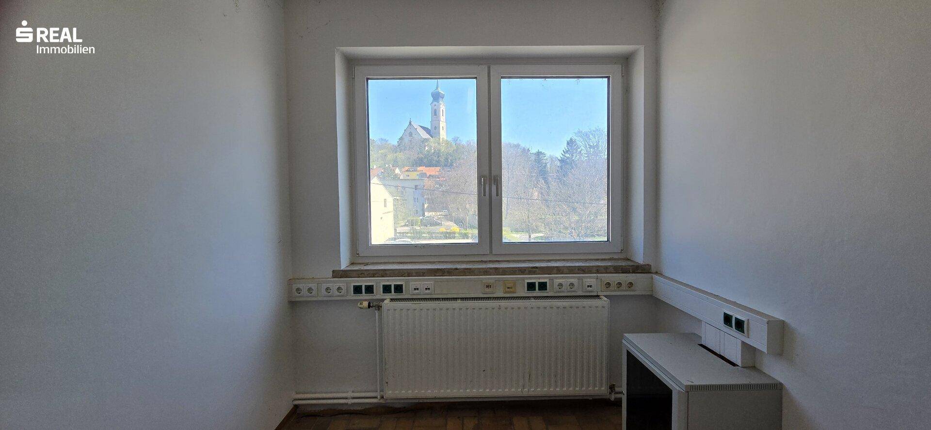 Helles Zimmer mit Parkettboden und einem Fenster, das einen Blick auf eine Kirche auf einem Hügel bietet.