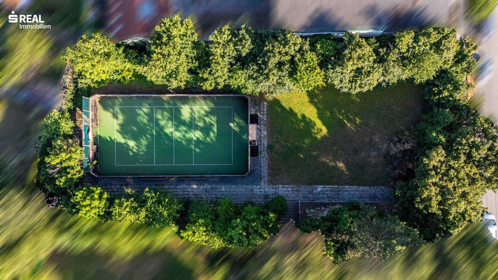 Draufsicht auf den privaten Tennisplatz, umgeben von dichtem Baumbestand und Rasenflächen.