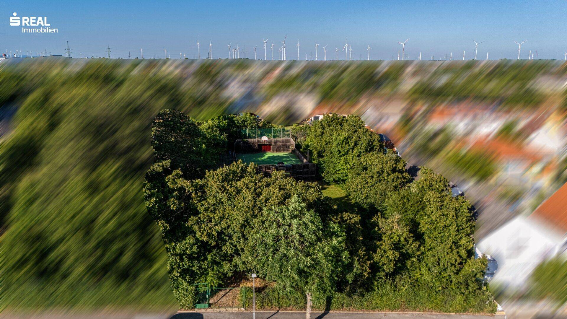 Luftaufnahme des Anwesens mit Tennisplatz, umgeben von Bäumen und Windrädern am Horizont.