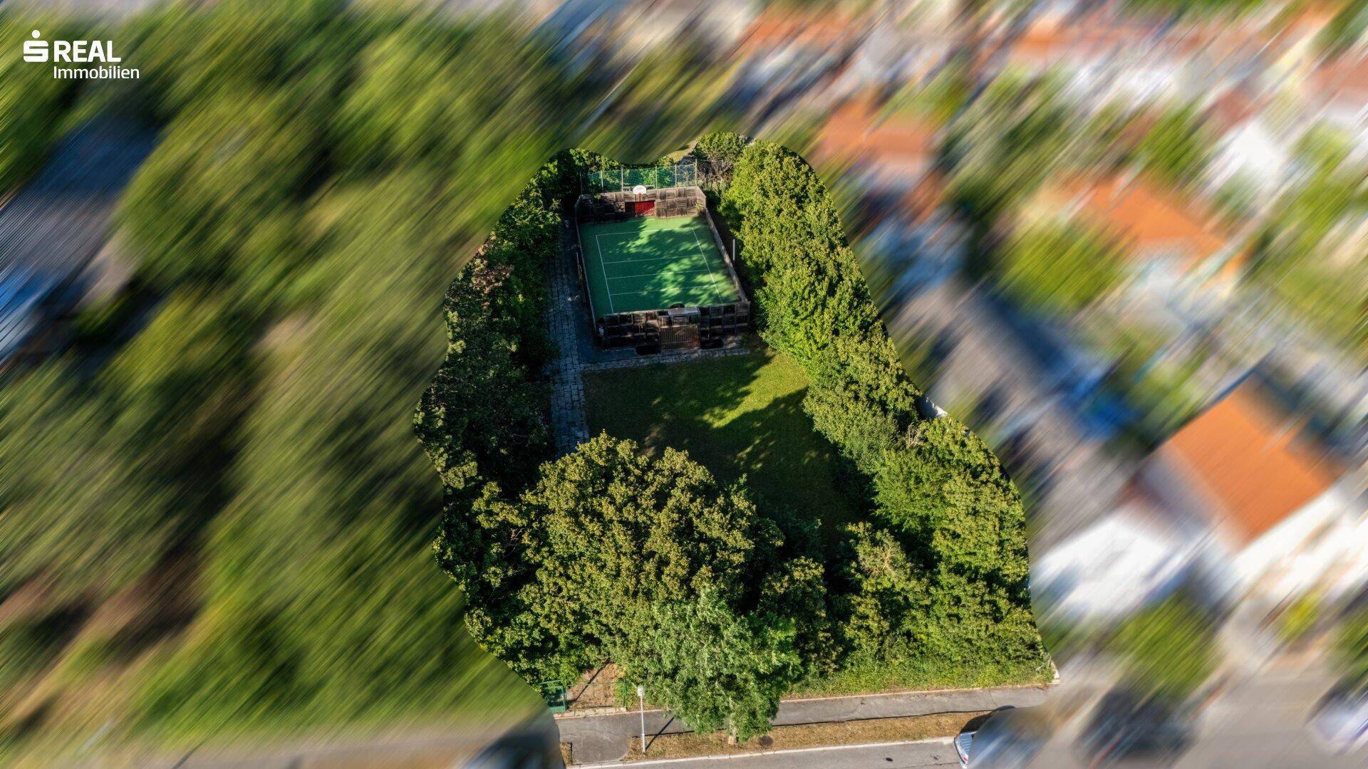 Weitläufige Luftaufnahme des Grundstücks mit Tennisplatz, eingebettet in eine grüne Landschaft.