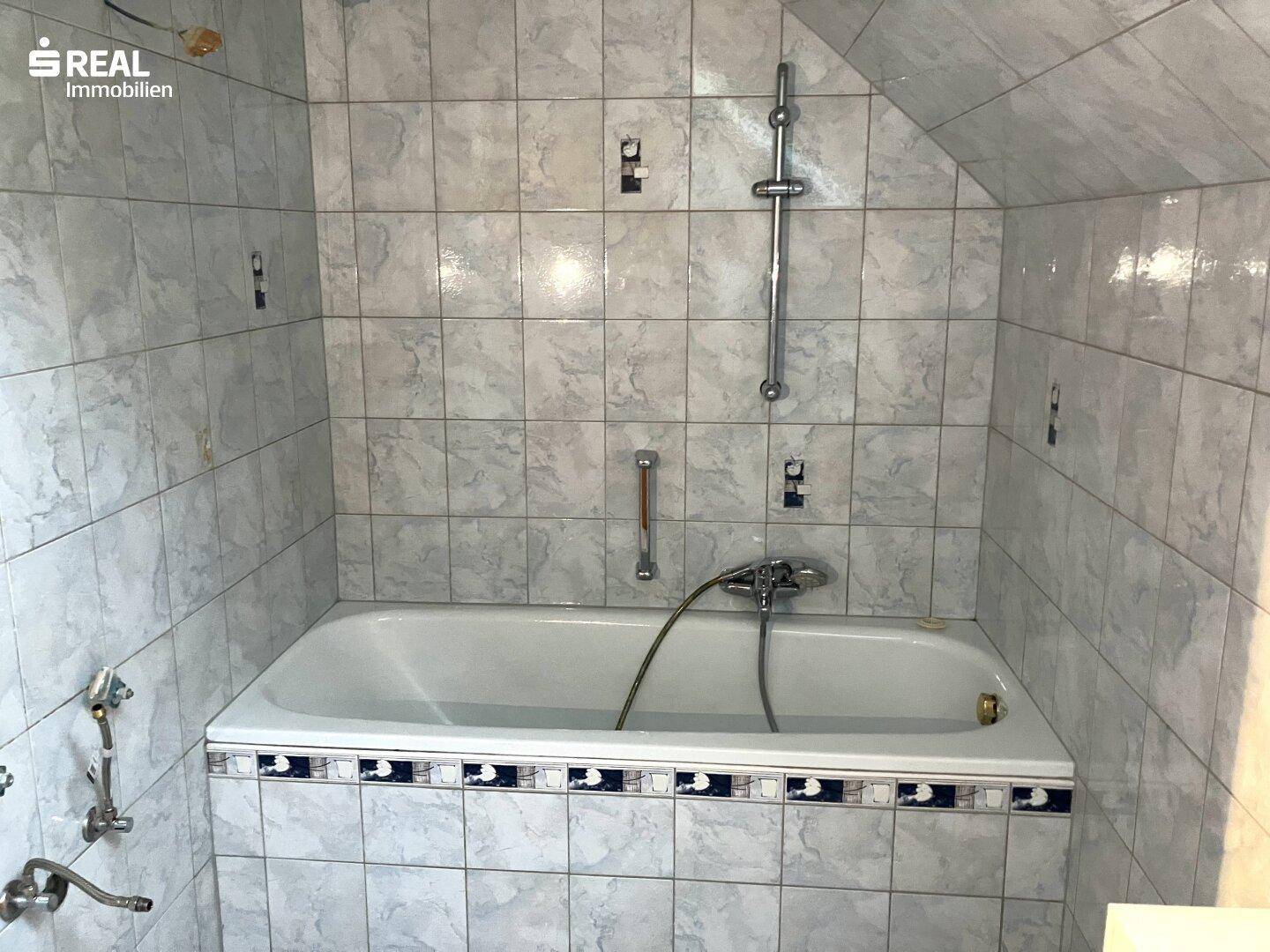 Badezimmer mit einer Badewanne und hellgrauen Fliesen, die eine Modernisierung vertragen könnte.