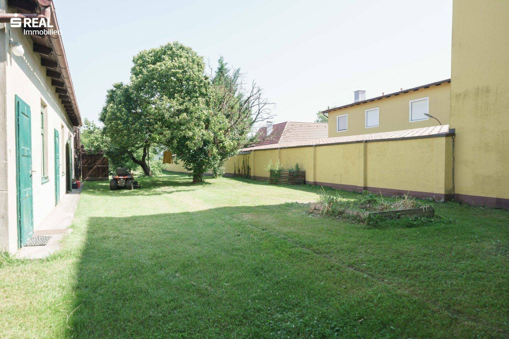 Weitläufiger Garten mit Rasenfläche zwischen zwei Gebäuden und einer hohen gelben Mauer.