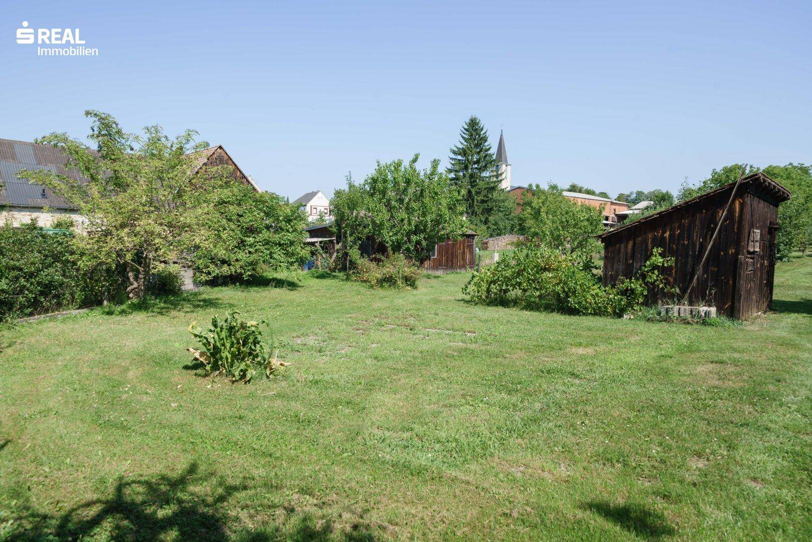 Großer, sonniger Garten mit Rasenfläche, Obstbäumen und einem rustikalen Holzschuppen im Hintergrund.