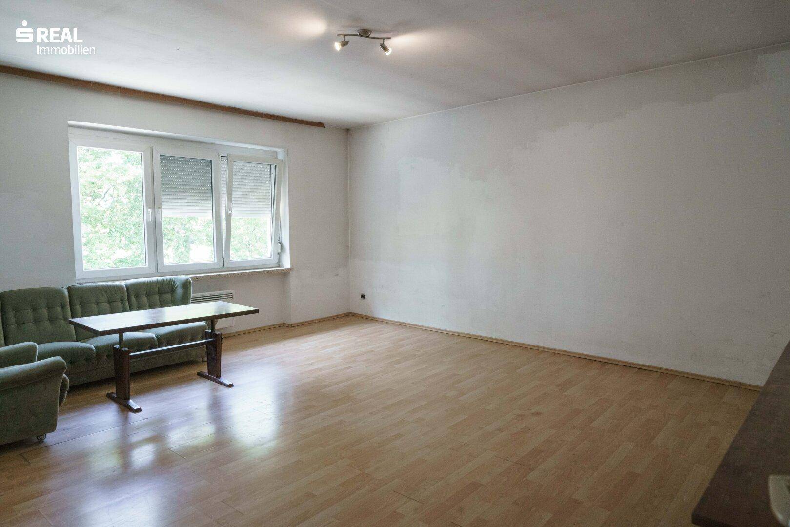 Geräumiges Wohnzimmer mit Laminatboden, großen Fenstern und einfacher Einrichtung.