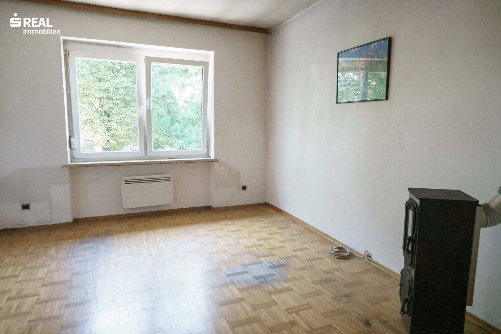 Zimmer mit Laminatboden, großem Fenster und einem Holzofen, der für Wärme sorgt.