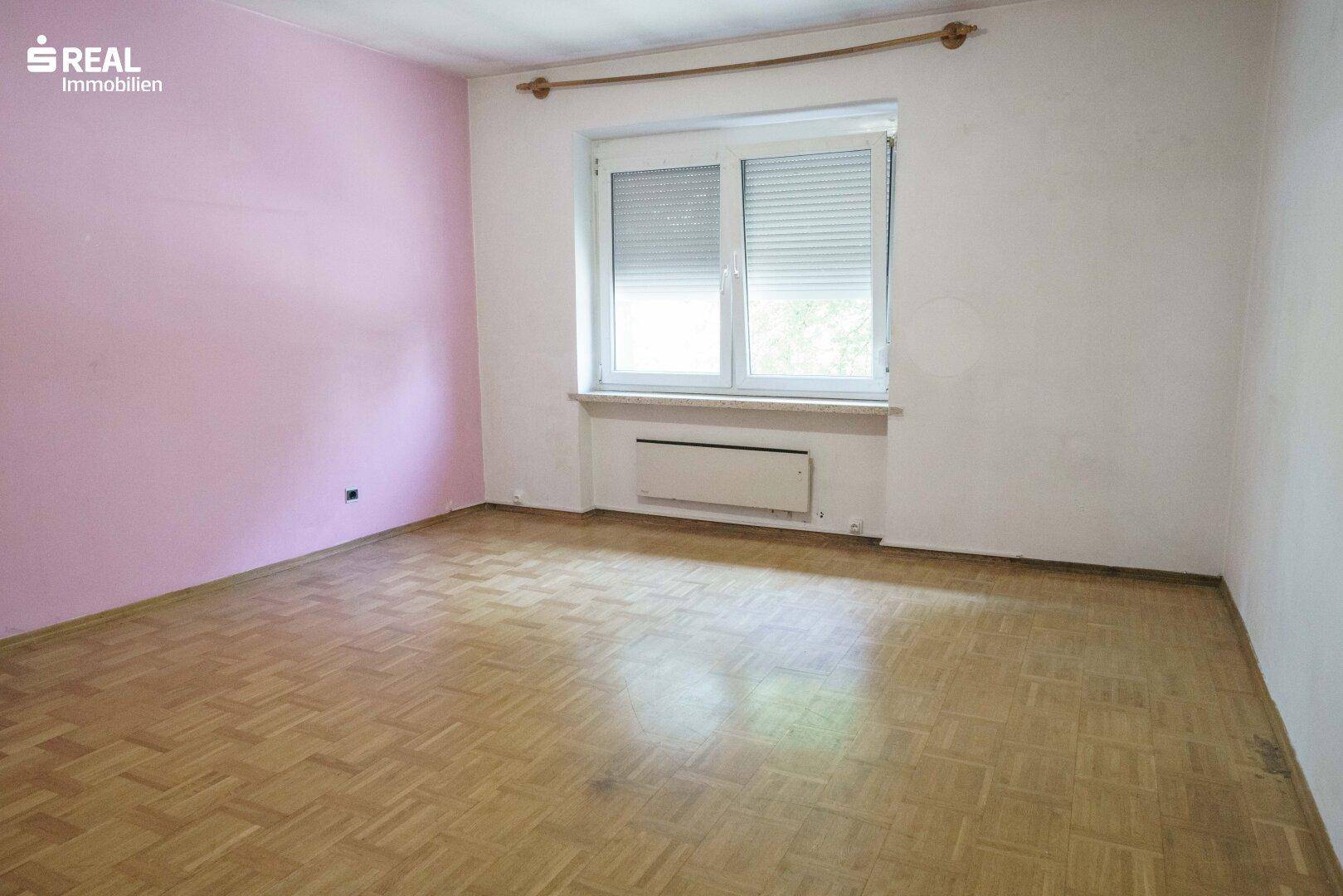 Schlafzimmer mit Laminatboden, Fenster und einer rosa Akzentwand, renovierungsbedürftig.
