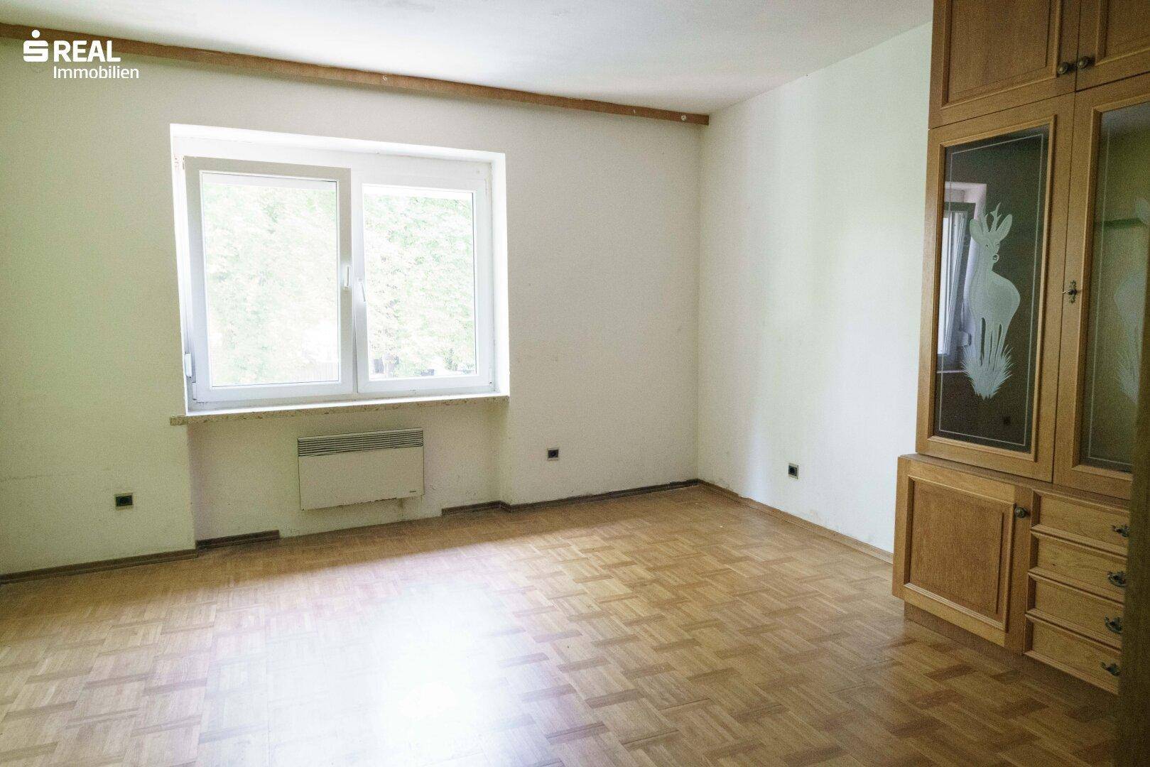 Geräumiges Zimmer mit Laminatboden, großem Fenster und einem Holzschrank.
