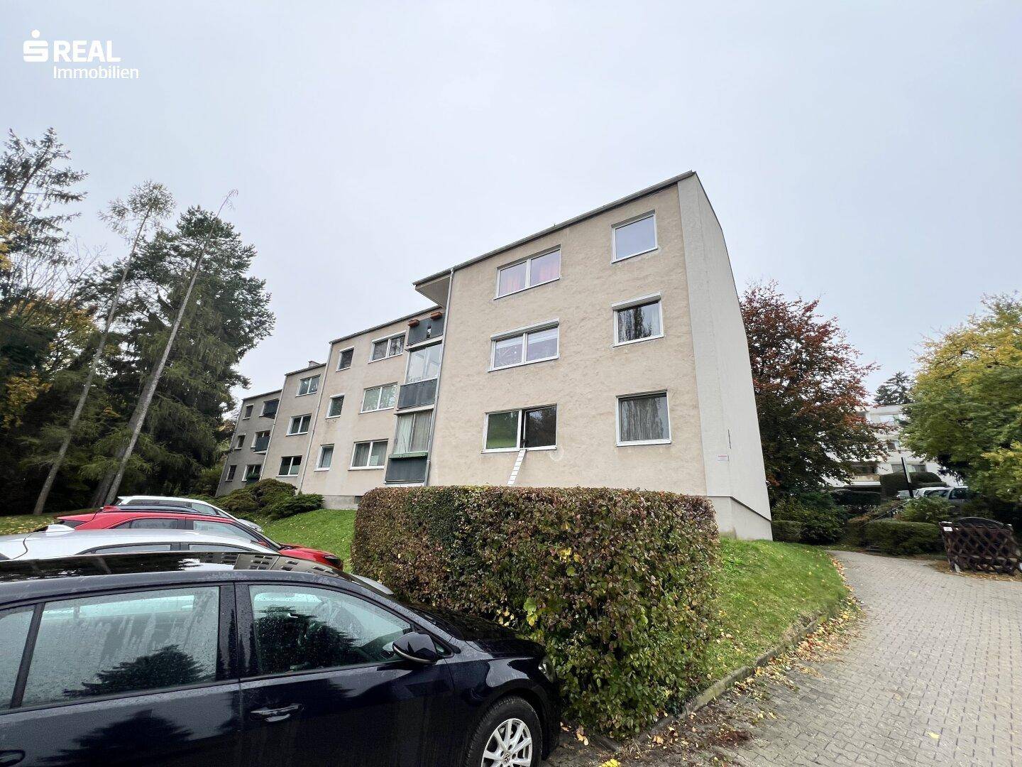 Mehrfamilienhaus mit Parkplätzen und einer Hecke im Vordergrund.