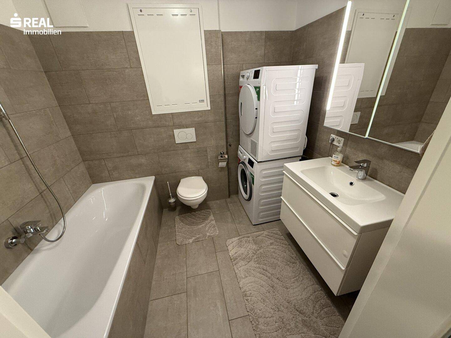 Modernes Badezimmer mit Badewanne, Waschbecken, WC und Waschmaschinenanschluss.