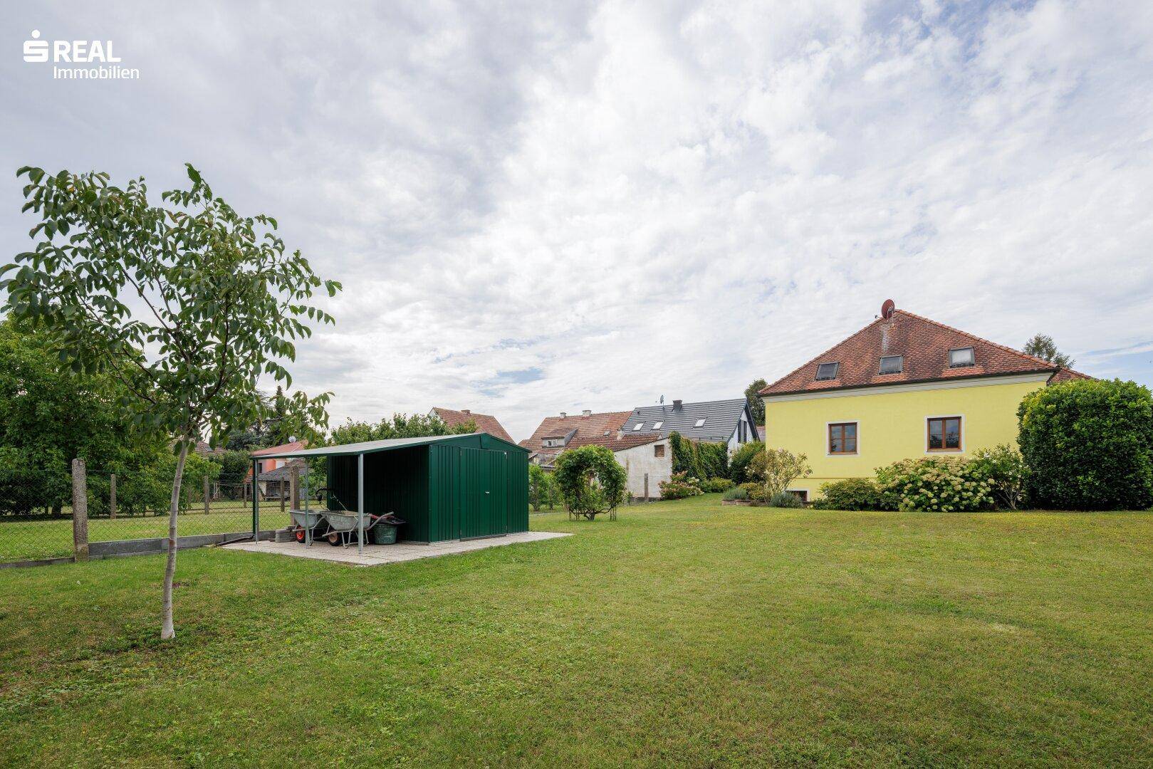 Weitläufiger, grüner Gartenbereich mit einem praktischen Geräteschuppen im Hintergrund und altem Baumbestand.