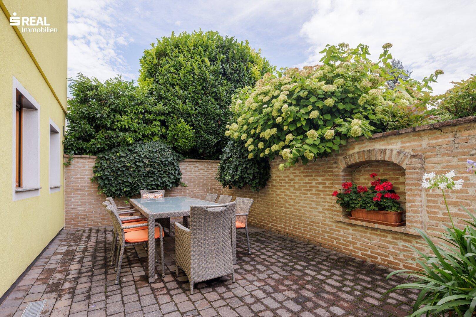 Gemütliche gepflasterte Terrasse mit Rattanmöbeln und üppiger Bepflanzung, ideal zum Entspannen.