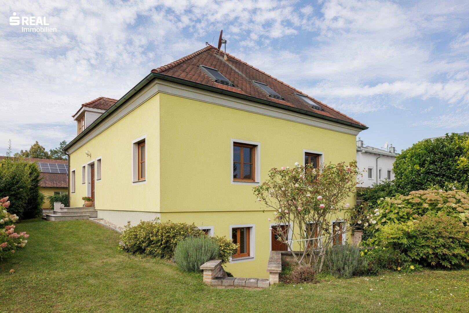 Seitenansicht des gelben Hauses mit Terrassenzugang und einem leicht abfallenden, grünen Gartenbereich.