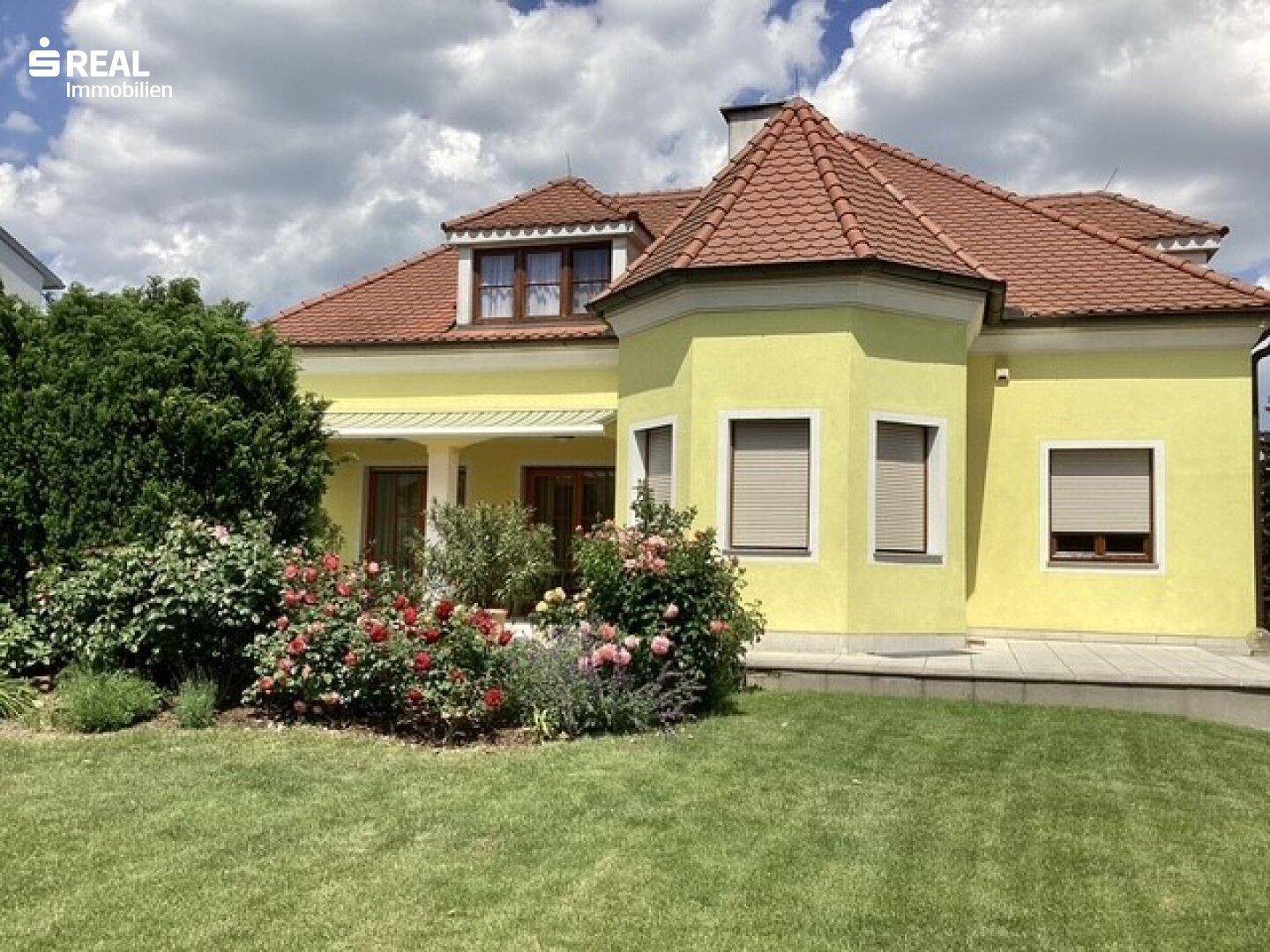 Ansicht des Hauses mit geschlossenen Rollläden, umgeben von einem blühenden Rosengarten und grüner Rasenfläche.