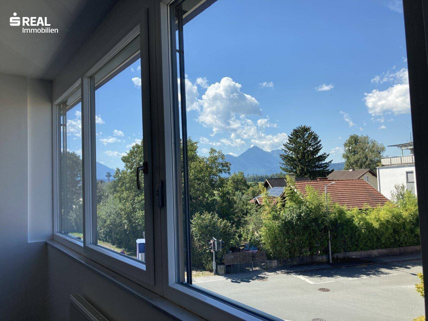 Weitblick aus dem Fenster auf grüne Bäume und majestätische Berge unter blauem Himmel.