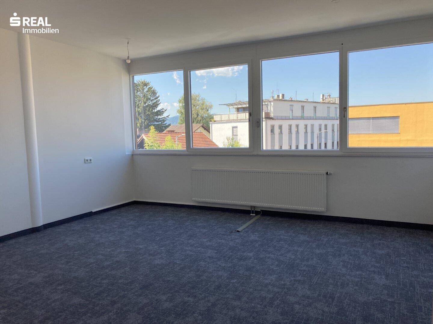 Heller Büroraum mit Teppichboden und großen Fenstern, die viel Tageslicht hereinlassen und eine weite Aussicht bieten.