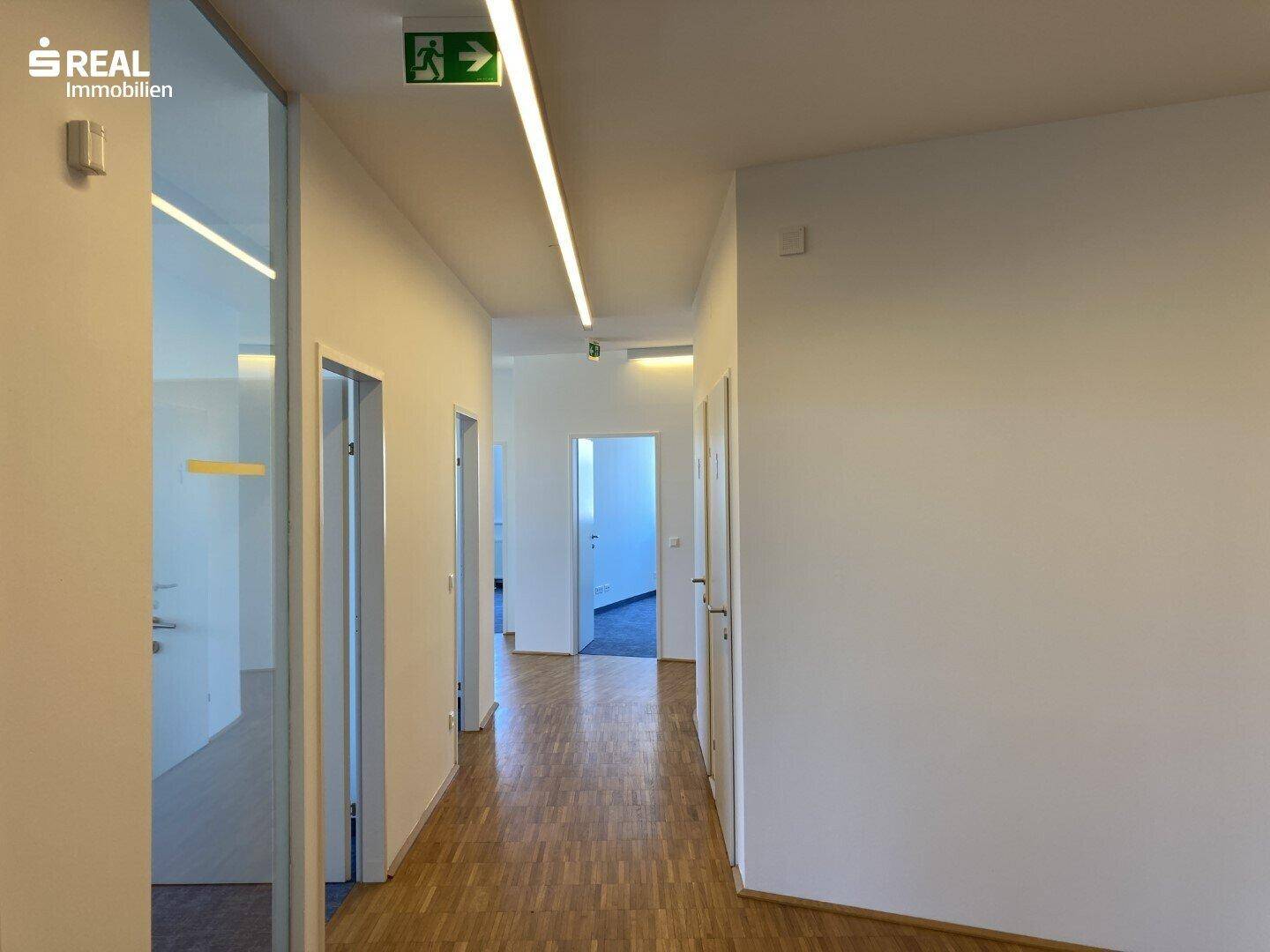 Langer Flur mit Holzparkettboden und modernen Deckenleuchten, führt zu mehreren Büroräumen.
