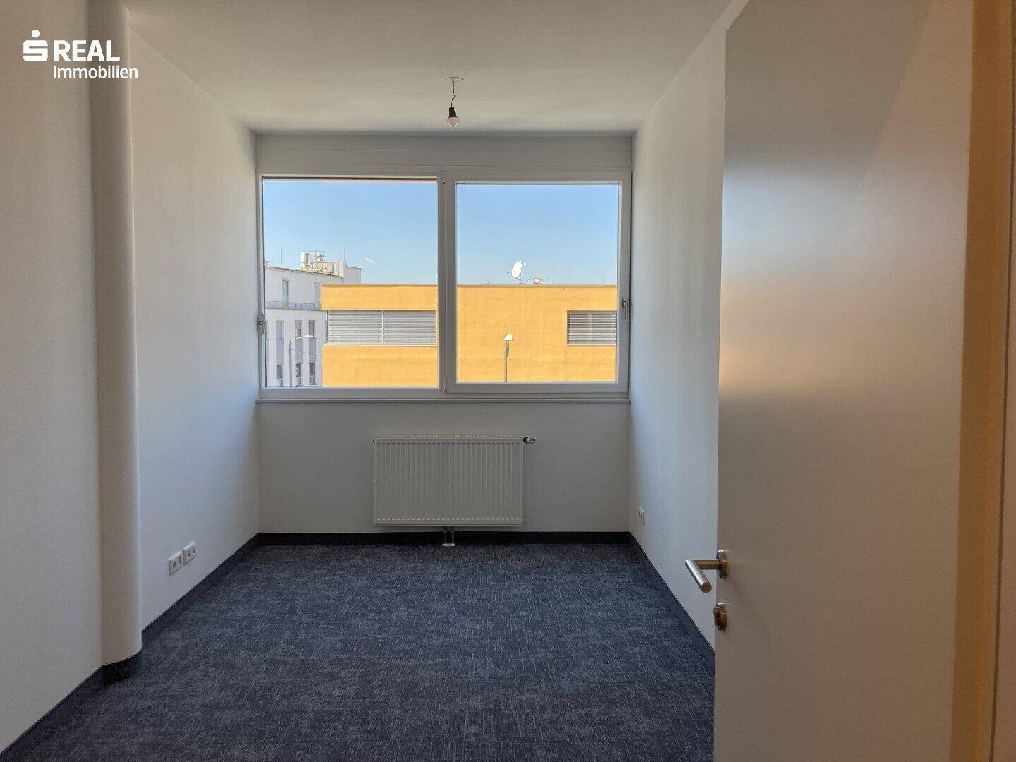 Kleiner, heller Büroraum mit Teppichboden und einem großen Fenster, das viel Tageslicht spendet.