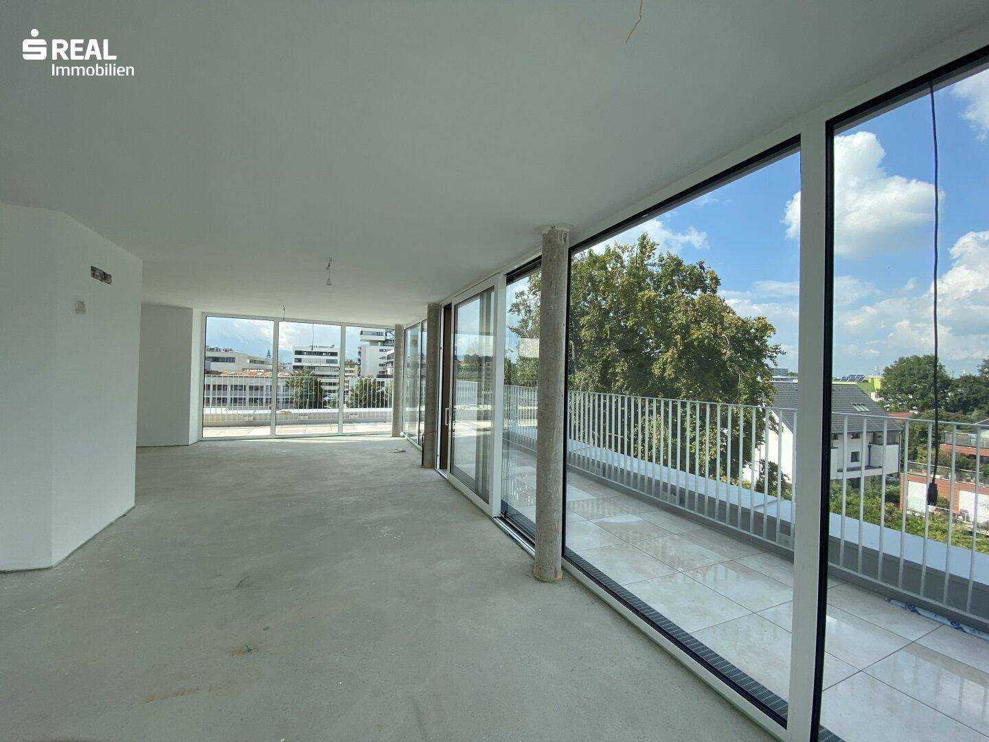 Weitläufiger Wohnbereich im Rohbau mit Panoramafenstern und Zugang zu einem umlaufenden Balkon mit Ausblick.