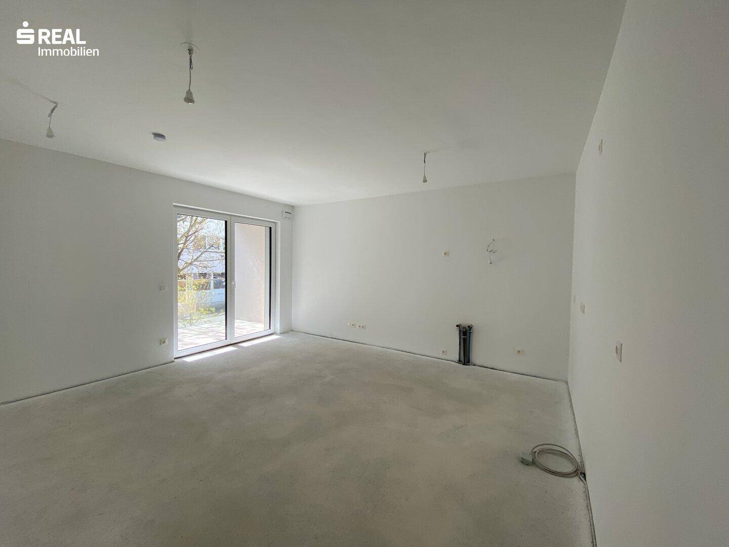 Leerer, heller Raum mit Rohbauzustand, großen Fenstern und Betonboden.