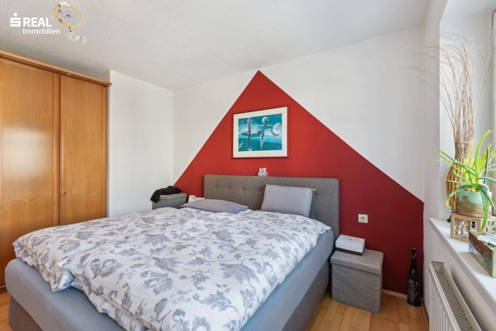 Schlafzimmer mit Doppelbett, einem Kleiderschrank und einer auffälligen roten Akzentwand, die dem Raum Charakter verleiht.
