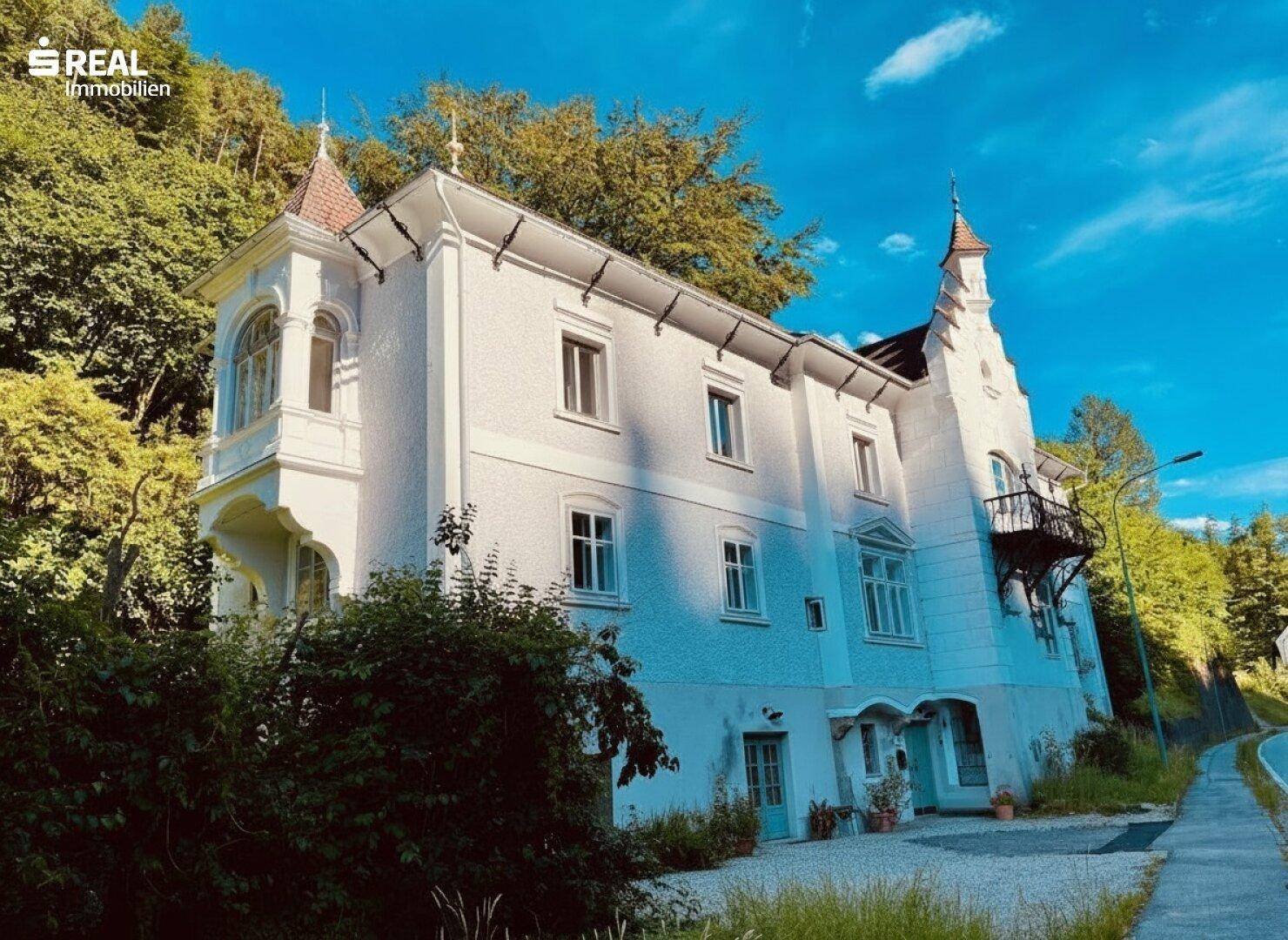 Seitenansicht des historischen Gebäudes mit Erker und großen Fenstern, umgeben von üppigem Grün.