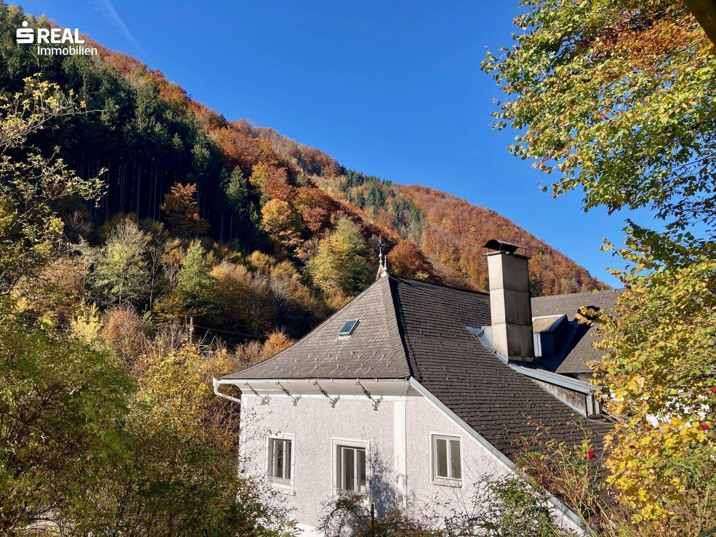 Historisches Gebäude umgeben von herbstlicher Natur, mit einem malerischen Blick auf die bewaldeten Berge.