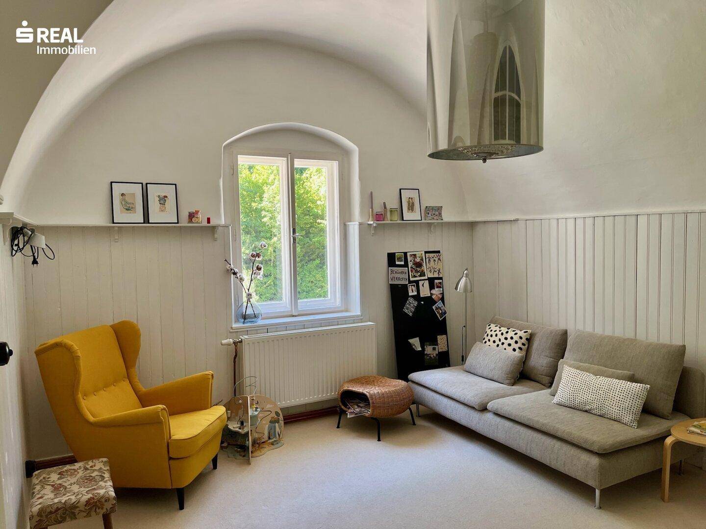 Helles Wohnzimmer mit gewölbter Decke, einem gelben Sessel und einem Sofa, mit Blick ins Grüne.