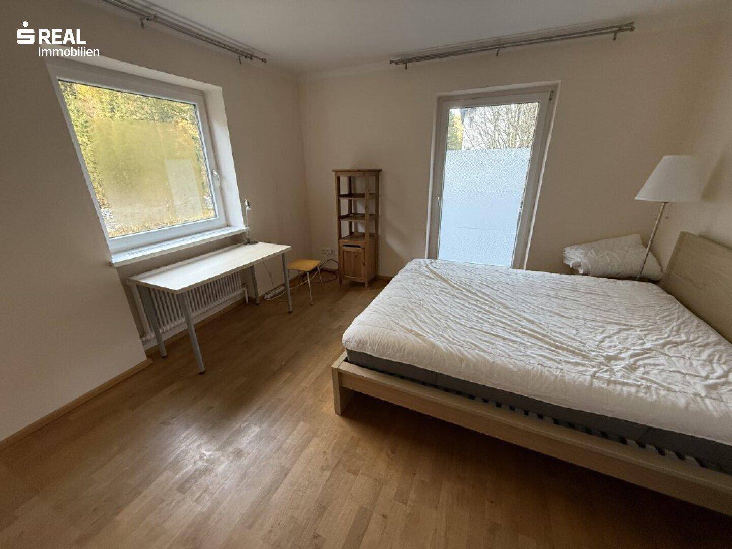 Schlafzimmer mit Holzboden, Doppelbett und zwei Fenstern, eines davon mit Sichtschutzfolie.