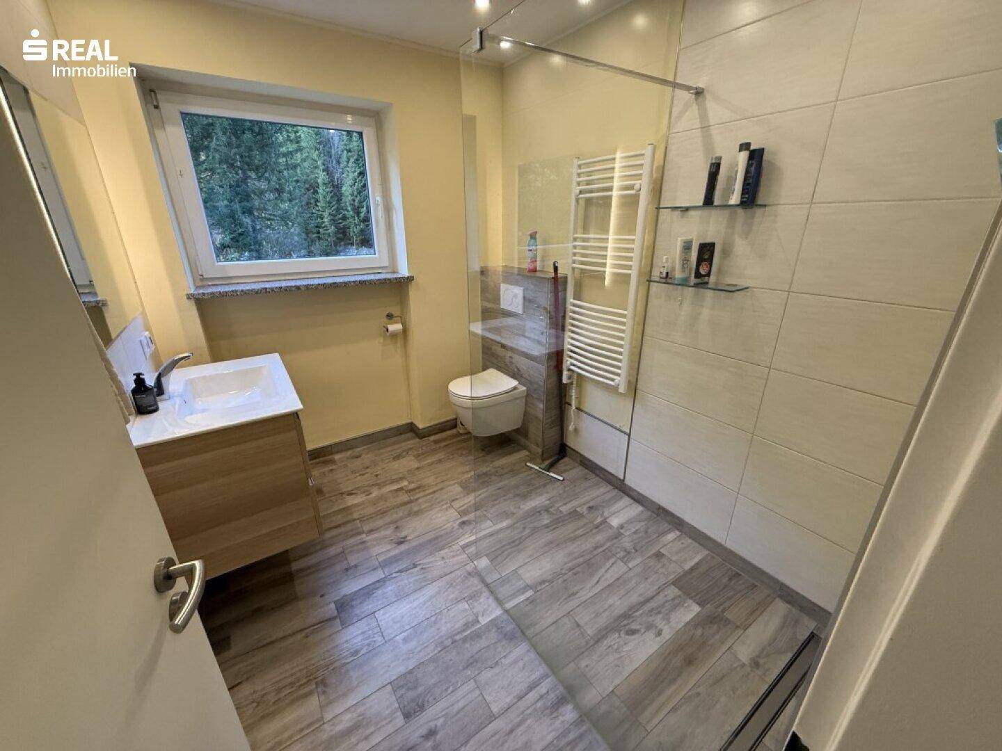 Modernes Badezimmer mit ebenerdiger Dusche, WC, Waschtisch und einem Fenster mit Waldblick.
