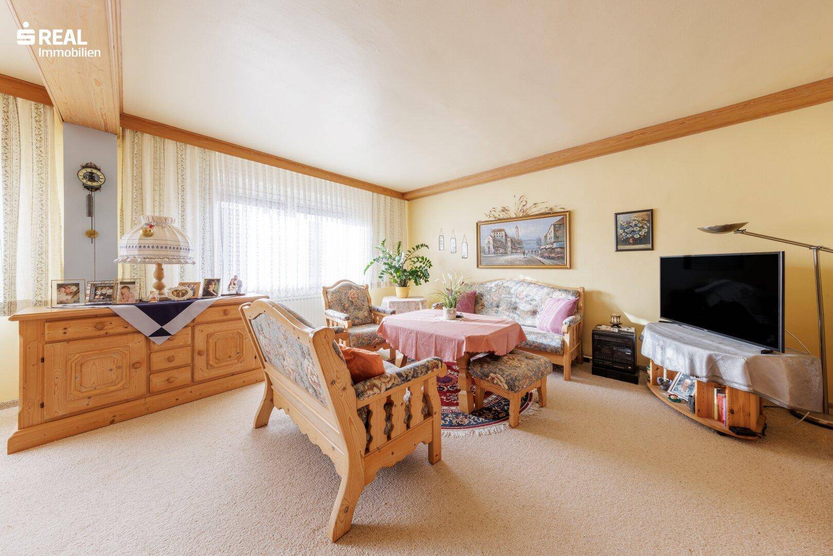 Helles Wohnzimmer mit Teppichboden, Holzbalken an der Decke und traditionellen Sitzmöbeln.