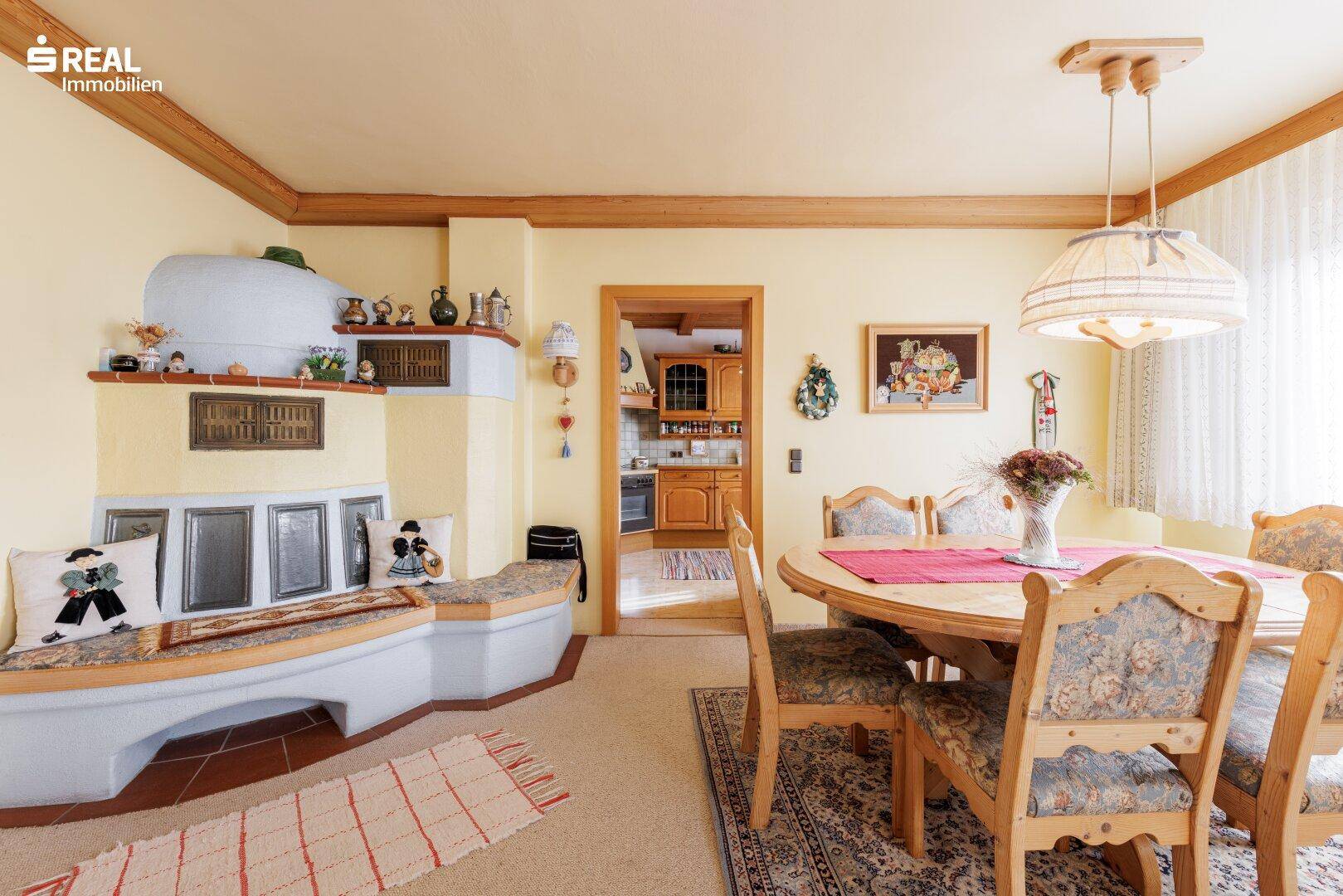 Esszimmer mit traditionellem Kachelofen, Teppichboden und Holzbalken, angrenzend an die Küche.