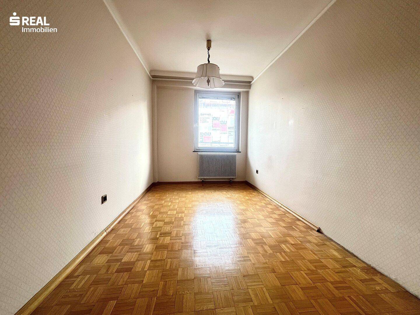 Leerer Raum mit Parkettboden und einem Fenster, das Tageslicht hereinlässt.