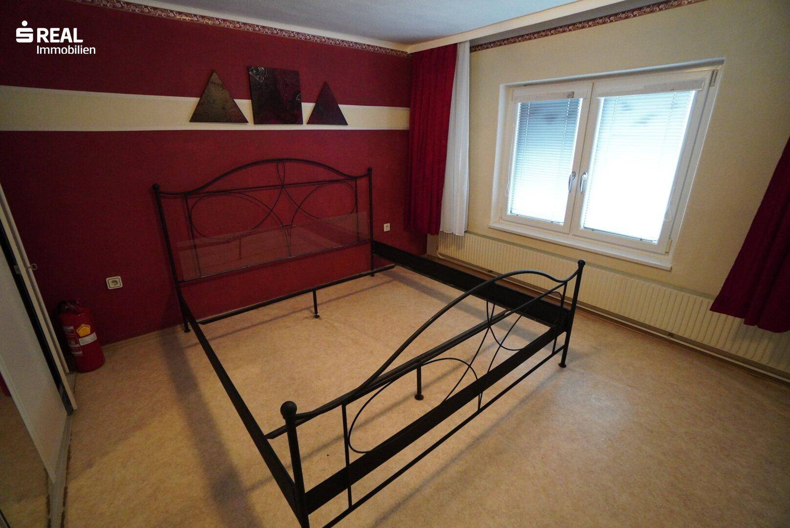Schlafzimmer mit roter Akzentwand, Metallbettrahmen und Fenster mit Jalousien.