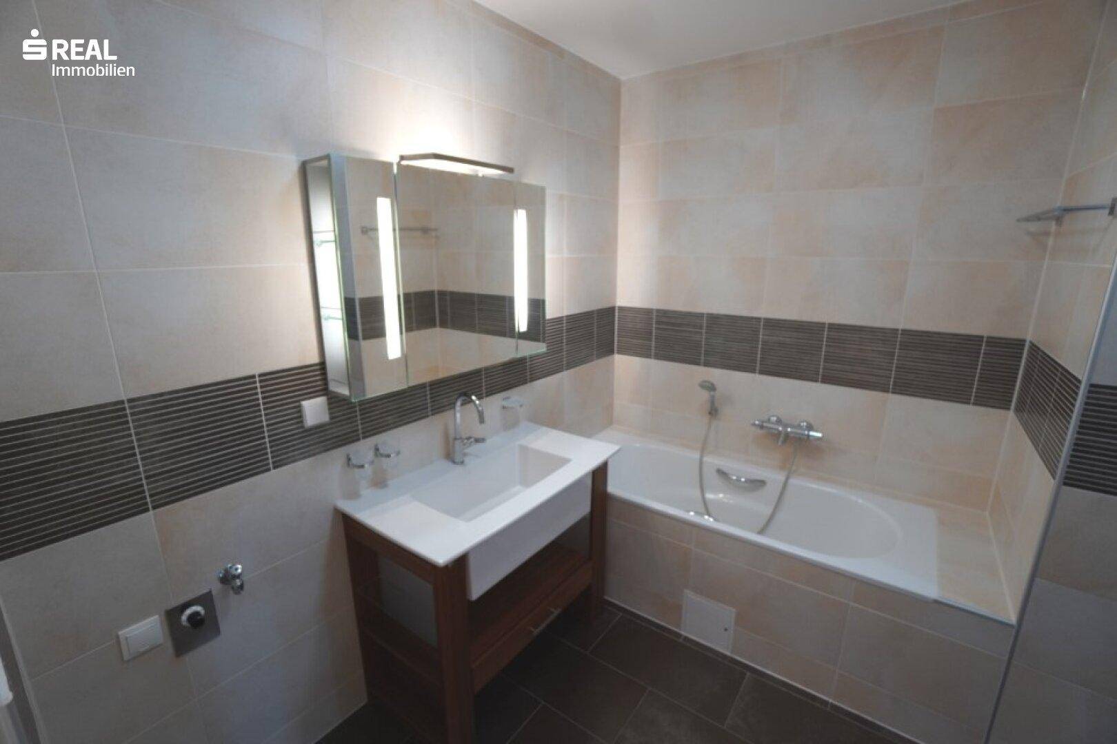 Modern gefliestes Badezimmer mit Badewanne, Waschtisch und beleuchtetem Spiegelschrank.