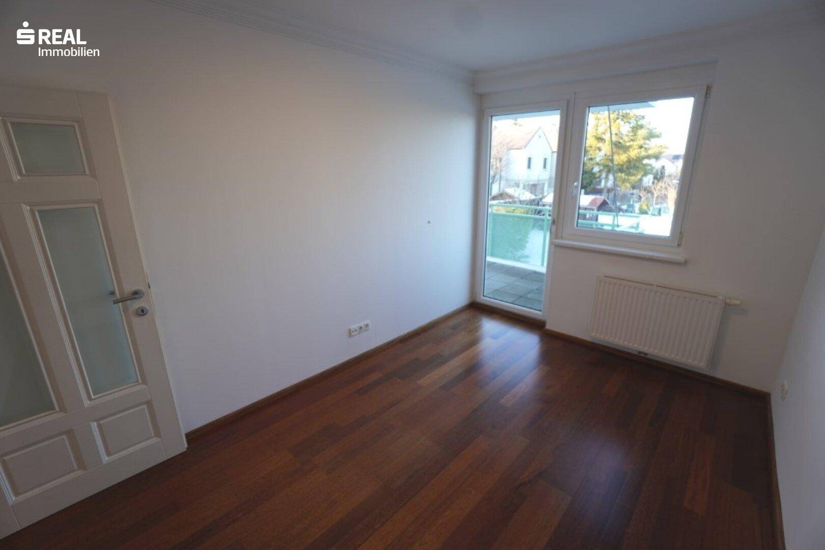 Helles Schlafzimmer mit dunklem Hartholzboden, großem Fenster und Zugang zum Balkon.