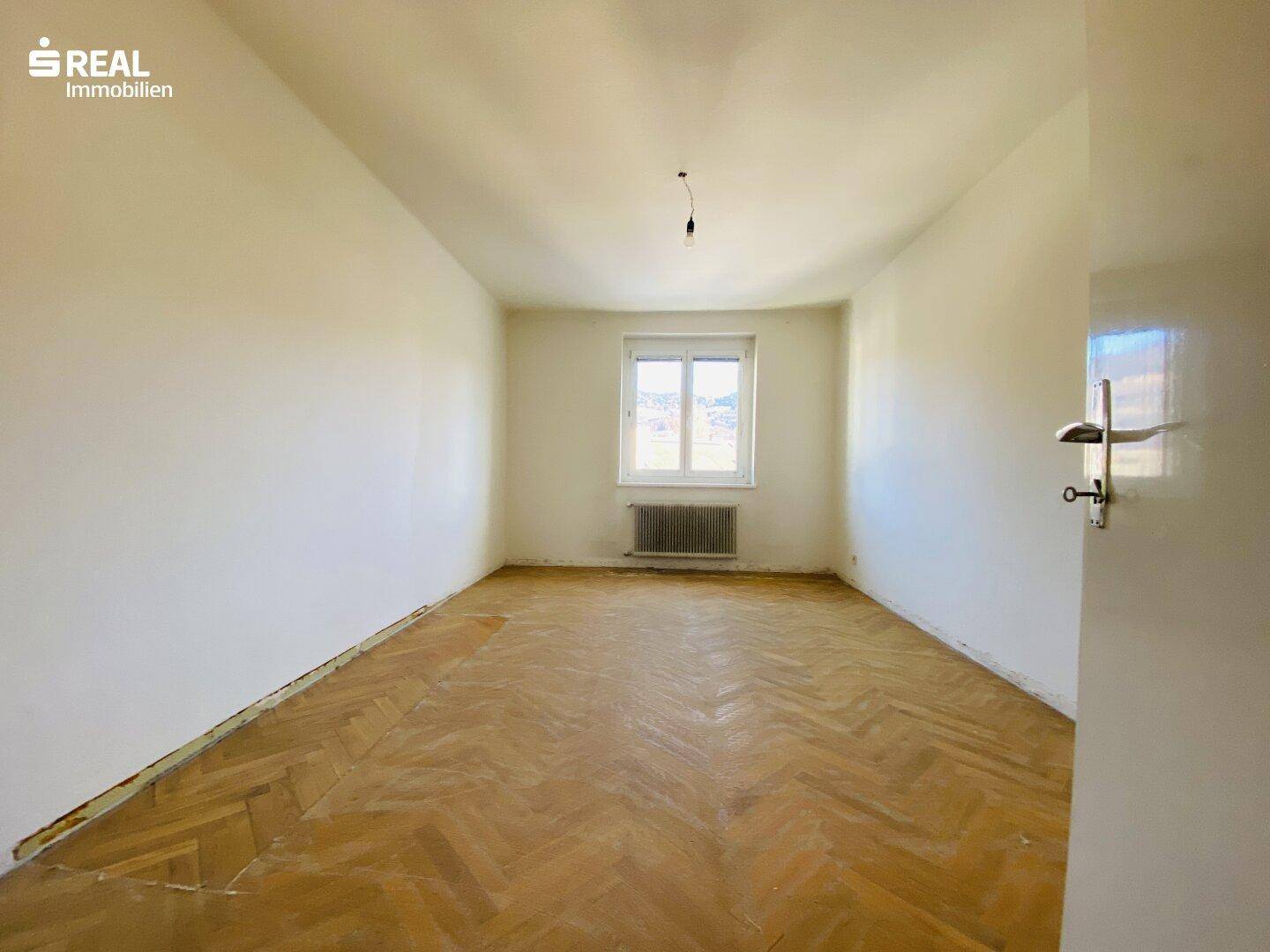 Leeres Zimmer mit Fischgrätparkett und einem Fenster, das viel Tageslicht hereinlässt und eine Renovierung erfordert.