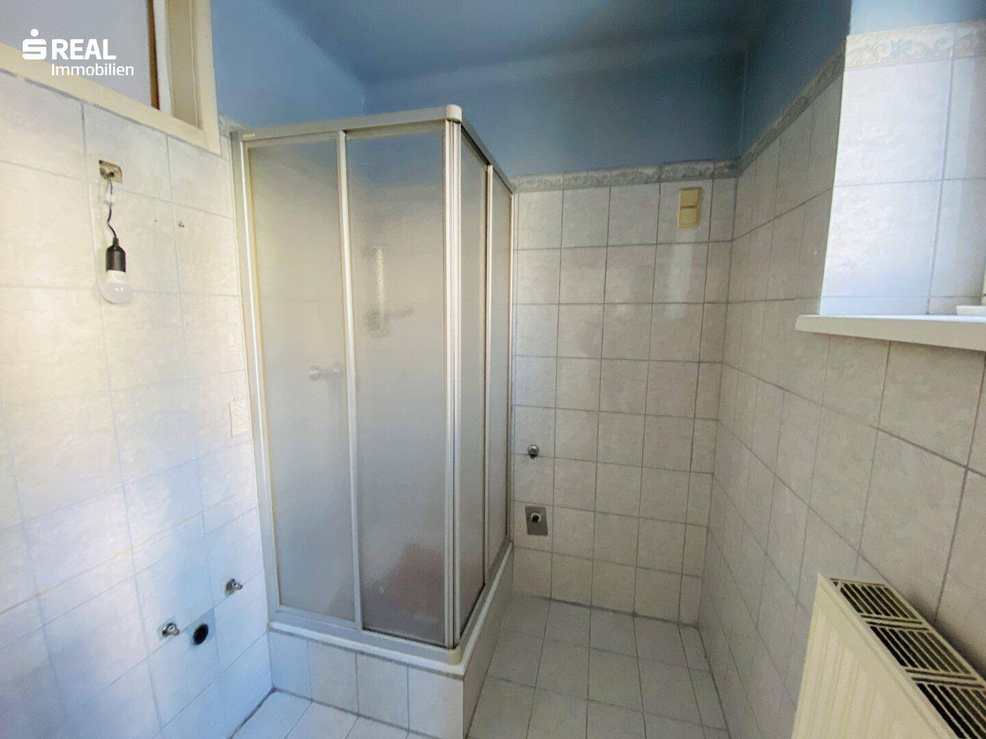 Badezimmer mit Duschkabine, weißen Fliesen und blauer Decke, renovierungsbedürftig.
